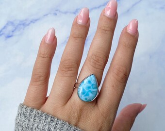 May include: A teardrop-shaped Larimar gemstone ring set in a polished silver band. The stone features a marbled pattern of sky blue and white tones, resembling clouds or ocean waves. The ring is worn on a finger, showcasing the smooth, polished surface.