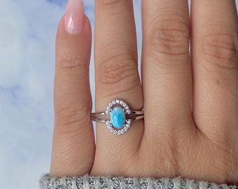 May include: A silver ring featuring a light blue oval gemstone, surrounded by a halo of small, clear stones. The ring has a split shank design. The ring is shown on a hand with a light pink manicure, against a light blue and white background.