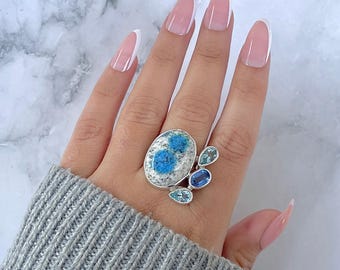 May include: A silver ring featuring an oval-shaped stone with blue and white patterns, accompanied by smaller blue gemstones. The ring is worn on a hand with a gray sweater.