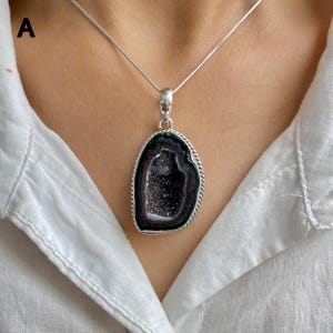 May include: A silver necklace featuring a large, irregular pendant with a dark, crystalline geode center. The stone is framed by a twisted silver border and suspended from a thin, smooth silver chain, resting against a white collared shirt.