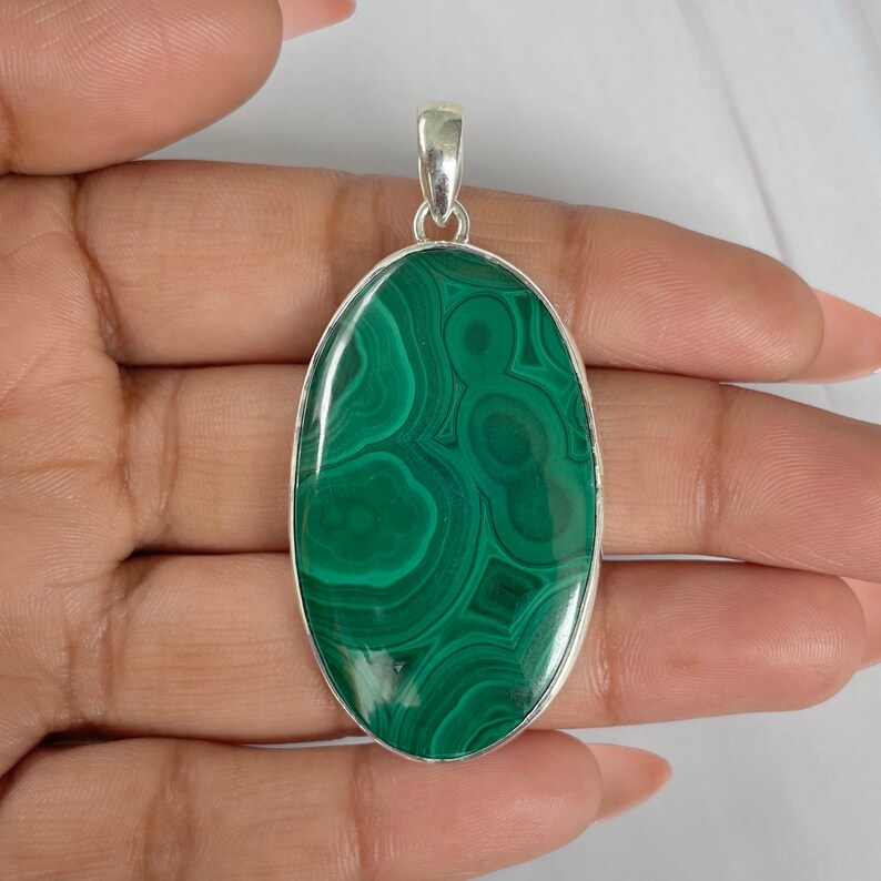Peut inclure: Pendentif ovale en malachite avec un b&eacute;li&egrave;re argent&eacute;. La pierre verte polie pr&eacute;sente un motif tourbillonnant de teintes vertes claires et fonc&eacute;es. Le pendentif mesure environ 5 cm de long.