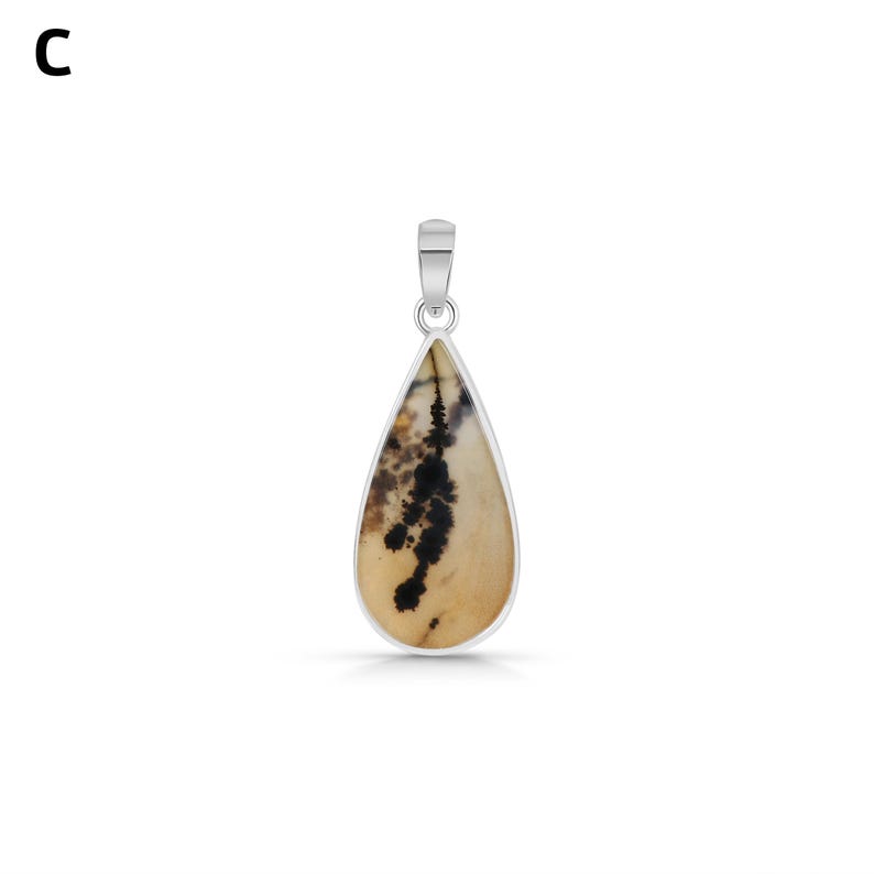 Puede incluir: Un colgante en forma de l&aacute;grima con una piedra beige e inclusiones dendr&iacute;ticas negras, engastado en un marco de metal plateado con una anilla a juego. La letra C es visible en la esquina superior izquierda de la imagen.