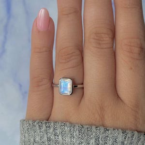 May include: A silver ring featuring a rectangular, iridescent gemstone. The gemstone displays light blue and white hues. The ring is worn on a finger, with light pink nail polish. The ring is a piece of jewellery.