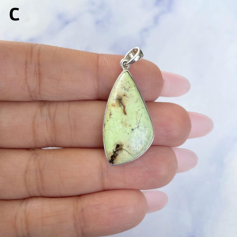 Pu&ograve; includere: Un ciondolo a forma di goccia con una pietra verde pallido e inclusioni di matrice marrone scuro, incastonato in una montatura in metallo color argento lucido con un piccolo occhiello circolare sulla parte superiore per una catenina.