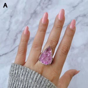 May include: A silver ring featuring a large, teardrop-shaped, pink gemstone. The gemstone has a textured, crystalline appearance. The ring is worn on a hand with pink nail polish and a gray sweater.