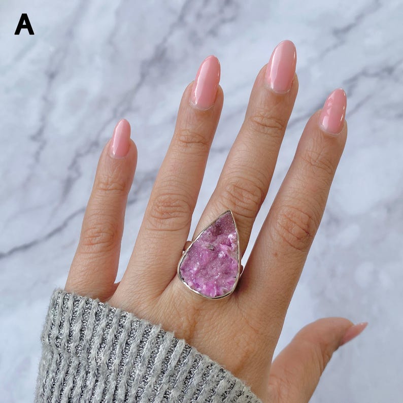 May include: A silver ring featuring a large, teardrop-shaped, pink gemstone. The gemstone has a textured, crystalline appearance. The ring is worn on a hand with pink nail polish and a gray sweater.