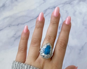Puede incluir: Un anillo de plata con una piedra grande en forma de lágrima con un patrón azul y blanco. El anillo se lleva en una mano con esmalte de uñas rosa. La letra "A" es visible en la esquina superior izquierda.