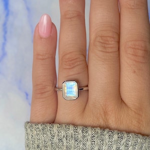 May include: A silver ring with a rectangular, iridescent gemstone. The gemstone displays light blue and white hues. The ring is set in a simple bezel. The ring is on a finger.