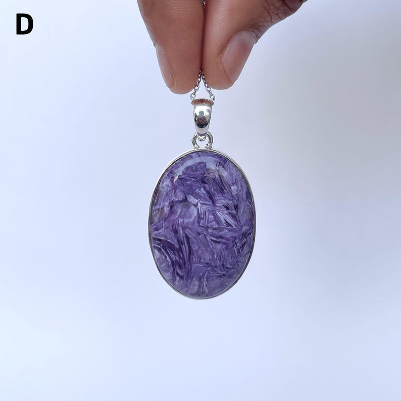 Op de afbeelding: Een ovaalvormige charoiet hanger met een wervelend patroon van dieppaarse en witte minerale insluitingen. De steen is gezet in een gepolijste zilverkleurige metalen rand met een bijpassend zilverkleurig oogje aan een dunne zilverkleurige ketting.