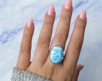 May include: A silver ring featuring a large, oval-shaped, light blue Larimar gemstone with white marbled patterns. The stone is set in a smooth, polished metal bezel, creating a clean and simple look that highlights the natural texture of the stone.