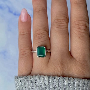 May include: A silver ring with a rectangular, emerald-green gemstone. The ring has a polished silver band. The gemstone is a vibrant green colour, and the ring is shown on a finger.