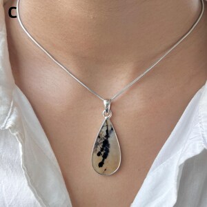 Puede incluir: Un collar de plata con un colgante en forma de l&aacute;grima de piedra lisa color crema con inclusiones dendr&iacute;ticas oscuras y ramificadas. El colgante est&aacute; sujeto por una sencilla anilla de plata a una cadena de serpiente fina y redonda.