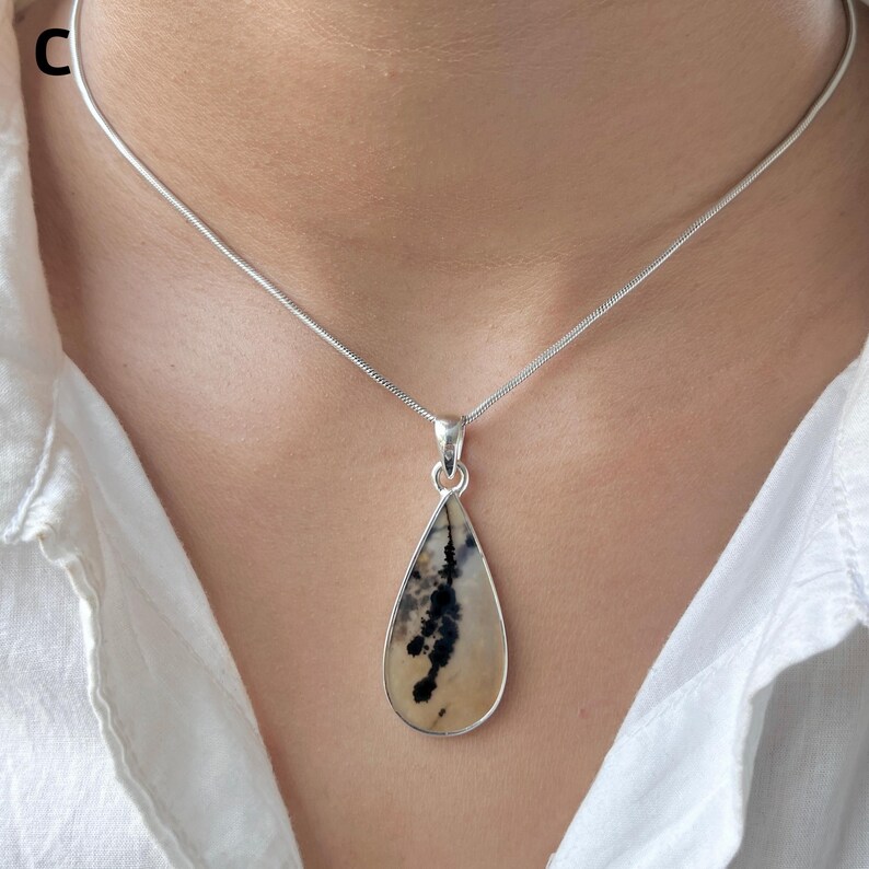 Puede incluir: Un collar de plata con un colgante en forma de l&aacute;grima de piedra lisa color crema con inclusiones dendr&iacute;ticas oscuras y ramificadas. El colgante est&aacute; sujeto por una sencilla anilla de plata a una cadena de serpiente fina y redonda.