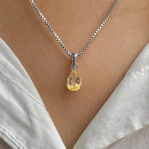 以下が含まれることがあります： 小さな黄色の宝石のペンダントが付いたシルバーのネックレス。ペンダントは繊細なチェーンから吊り下げられており、白いシャツの上に着用されています。