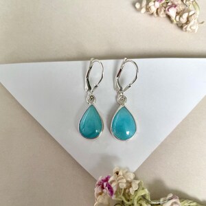 Puede incluir: Un par de pendientes de plata con cierre de palanca y piedras preciosas azul claro en forma de lágrima. Las piedras preciosas están engastadas en un bisel de plata. Los pendientes se exhiben sobre una superficie blanca con detalles florales.