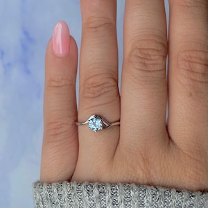 Könnte beinhalten: Ein silberner Ring mit einem runden, hellblauen Edelstein. Das Band des Rings hat ein geteiltes Design auf beiden Seiten des Edelsteins. Der Ring wird an einem Finger gezeigt.