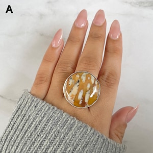 May include: A round ring featuring a polished stone with a mottled pattern of mustard yellow, cream, and dark brown spots. The stone is set in a smooth, thin silver-toned metal bezel that encircles the circular cabochon, creating a bold statement piece.