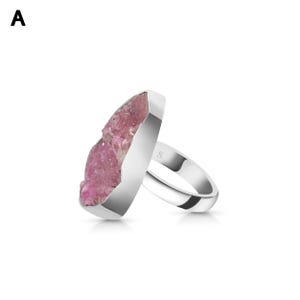 May include: A silver ring featuring a large, irregular-shaped pink crystal. The crystal has a rough, textured surface and is set in a polished silver band. The ring is displayed against a white background.
