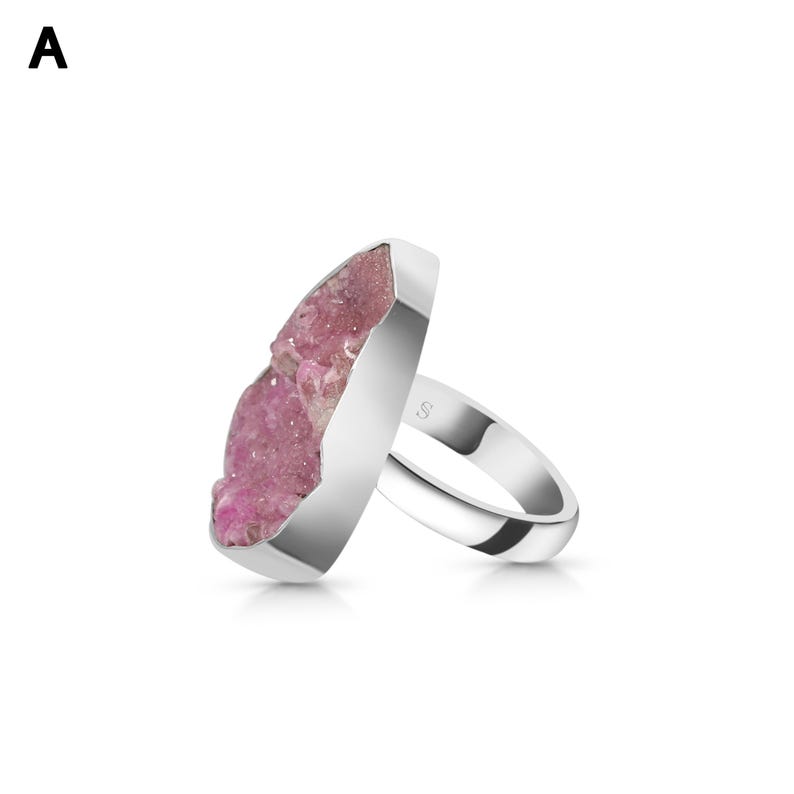 May include: A silver ring featuring a large, irregular-shaped pink crystal. The crystal has a rough, textured surface and is set in a polished silver band. The ring is displayed against a white background.