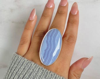 May include: A large, oval-shaped ring with a light blue stone and a silver band. The stone has a layered pattern of varying shades of blue. The ring is worn on a finger.