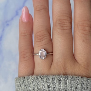 May include: A silver ring with an oval, light pink gemstone. The ring is set on a finger, with a light pink nail polish visible. The background is a blurred mix of blue and white.