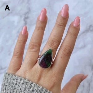 Puede incluir: Un anillo de plata con una gran piedra de zoisita en forma de lágrima, con inclusiones de color magenta intenso y verde oscuro. La piedra está engastada en un bisel de metal liso y la letra A aparece en la esquina superior izquierda.