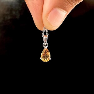以下が含まれることがあります： 洋梨型の、金色がかった黄色の宝石ペンダントが付いたシルバーネックレス。ペンダントはシルバーの台座にセットされ、繊細なチェーンから吊り下げられています。背景は黒です。