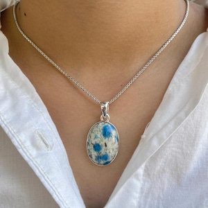Op de afbeelding: Een zilveren ketting met een ovale hanger in blauw en wit. De hanger is gemaakt van een natuursteen met een gevlekt patroon.