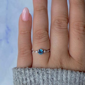 May include: A silver ring with a large, round, blue gemstone at the center, flanked by smaller, clear stones. The ring is set on a finger, showcasing its delicate design and sparkling details. The ring is a piece of jewelry.