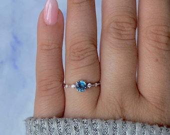 May include: A silver ring with a large, round, blue gemstone at the center, flanked by smaller, clear stones. The ring is set on a finger, showcasing its delicate design and sparkling details. The ring is a piece of jewelry.