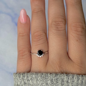 May include: A silver ring with a prominent, round black gemstone. The ring is embellished with smaller, clear gemstones arranged in a curved pattern below the black stone. The ring is shown on a finger.