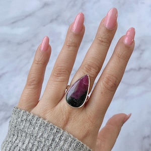 May include: A silver ring featuring a large, teardrop-shaped gemstone. The stone displays a vibrant mix of colors, with deep red and dark green hues. The ring is worn on a hand, showcasing the polished stone's unique pattern.