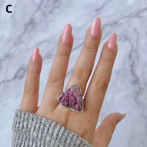 May include: A silver ring featuring a large, triangular, rough-cut pink gemstone. The ring's setting is a simple silver band, and the gemstone has a textured, natural appearance. The ring is shown on a hand with pink nail polish.