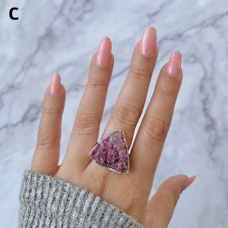 May include: A silver ring featuring a large, triangular, rough-cut pink gemstone. The ring's setting is a simple silver band, and the gemstone has a textured, natural appearance. The ring is shown on a hand with pink nail polish.