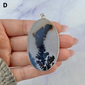 May include: An oval-shaped pendant featuring a translucent, milky white stone with dark, fern-like dendritic inclusions. The stone is encased in a polished silver-toned metal frame with a small bail at the top for hanging. The letter D is visible.