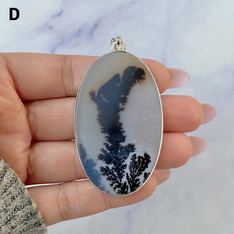 May include: An oval-shaped pendant featuring a translucent, milky white stone with dark, fern-like dendritic inclusions. The stone is encased in a polished silver-toned metal frame with a small bail at the top for hanging. The letter D is visible.