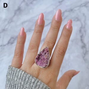 May include: A large, silver-toned ring featuring a teardrop-shaped, rough-cut pink gemstone. The gemstone has a textured, crystalline appearance. The ring is worn on a hand with pink nail polish and a gray sweater.