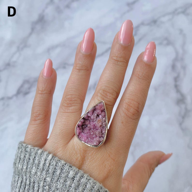 May include: A large, silver-toned ring featuring a teardrop-shaped, rough-cut pink gemstone. The gemstone has a textured, crystalline appearance. The ring is worn on a hand with pink nail polish and a gray sweater.