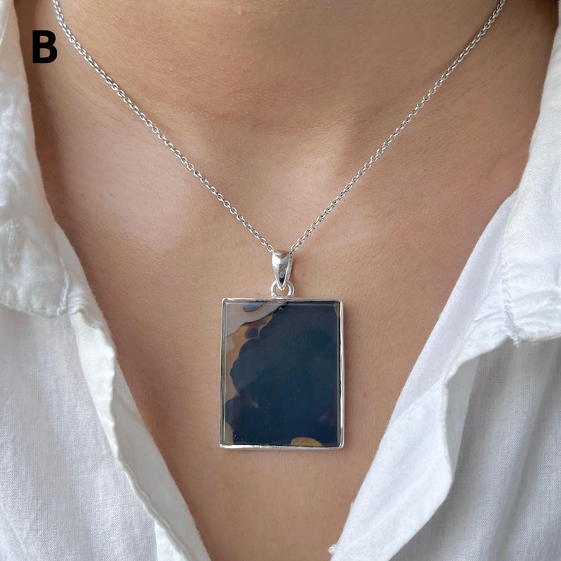 May include: A silver necklace featuring a rectangular pendant with a dark, polished stone inlay. The stone displays deep blue and black tones with subtle, lighter brown organic patterns. The pendant hangs from a thin, delicate silver chain.