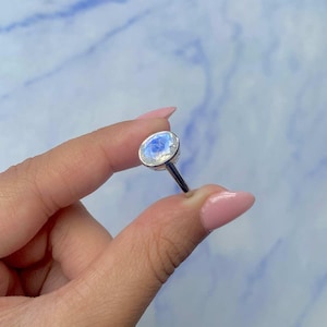 May include: A silver ring featuring a large, oval-cut moonstone. The moonstone has a milky, iridescent appearance with blue and white hues. The ring is held against a light blue and white background.