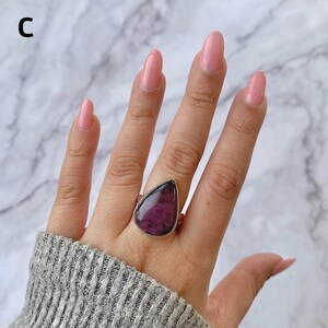 以下が含まれることがあります： 銀色の指輪で、深いマゼンタと黒の大きな涙型の宝石が滑らかな金属製の枠にはめ込まれています。画像の左上隅に文字Cが見えます。指輪は指にはめられており、石の暗い色合いが特徴的です。