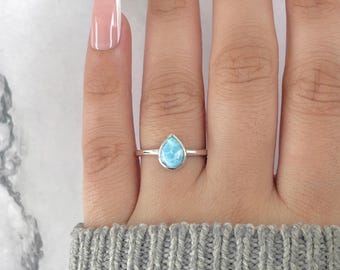 May include: A silver ring featuring a pear-shaped, light blue gemstone. The ring is set on a slender band and is displayed on a person's finger. The gemstone has a marbled appearance, and the ring is worn with a gray sweater.