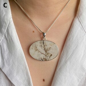 May include: A silver necklace featuring an oval-shaped pendant with a smooth, off-white stone surface. The stone displays natural, dark brown, tree-like dendritic inclusions that branch across the center. The pendant hangs from a thin silver chain.