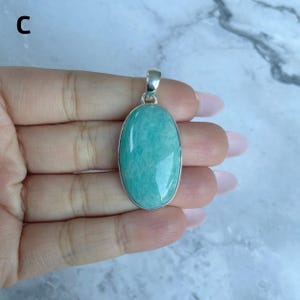 May include: An oval-shaped pendant featuring a smooth, polished light teal amazonite gemstone set in a simple silver-toned metal frame with a small bail at the top for attachment to a chain. The stone displays natural color variations.