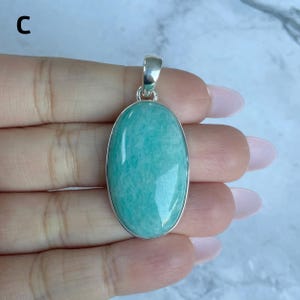 May include: An oval-shaped pendant featuring a smooth, polished light teal gemstone set in a simple silver-toned metal frame with a small bail at the top. The letter C is visible in the upper left corner of the image.