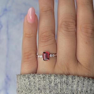 May include: A silver ring featuring a rectangular, deep red gemstone as the centerpiece, flanked by smaller, round, clear stones. The ring is set against a hand wearing a gray sweater, with a light blue background.