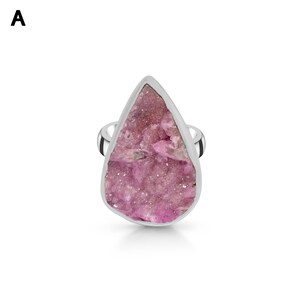 May include: A silver ring featuring a large, teardrop-shaped pink gemstone. The gemstone has a rough, textured surface and is set in a polished silver band. The ring is displayed against a white background.