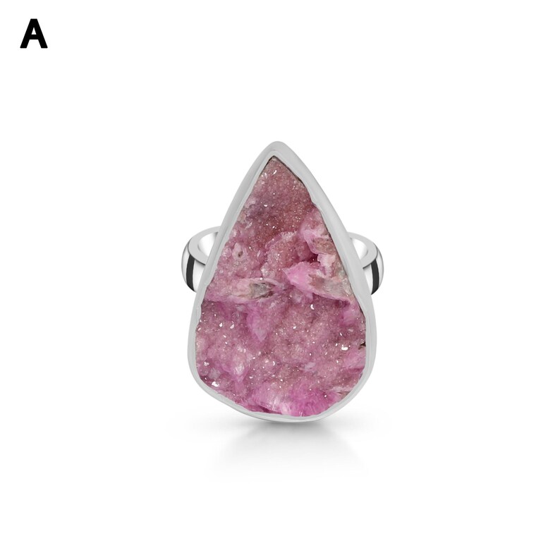 May include: A silver ring featuring a large, teardrop-shaped pink gemstone. The gemstone has a rough, textured surface and is set in a polished silver band. The ring is displayed against a white background.