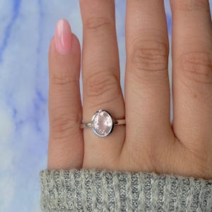 Op de afbeelding: Een zilveren ring met een grote, ovaalvormige, lichtroze edelsteen. De ring heeft een eenvoudige, gepolijste zilveren band. De edelsteen is het middelpunt en toont zijn zachte kleur en gladde oppervlak.