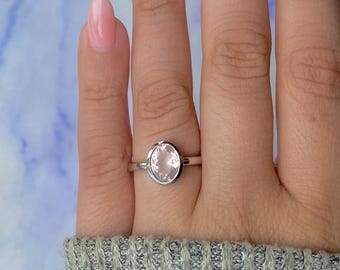 Puede incluir: Un anillo de plata con una gema grande, ovalada, de color rosa pálido. El anillo tiene una banda de plata pulida y sencilla. La gema es el punto focal, mostrando su color suave y superficie lisa.